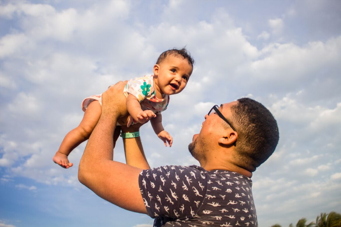 A delightful moment of a father lovingly lifting his happy baby against a clear sky.