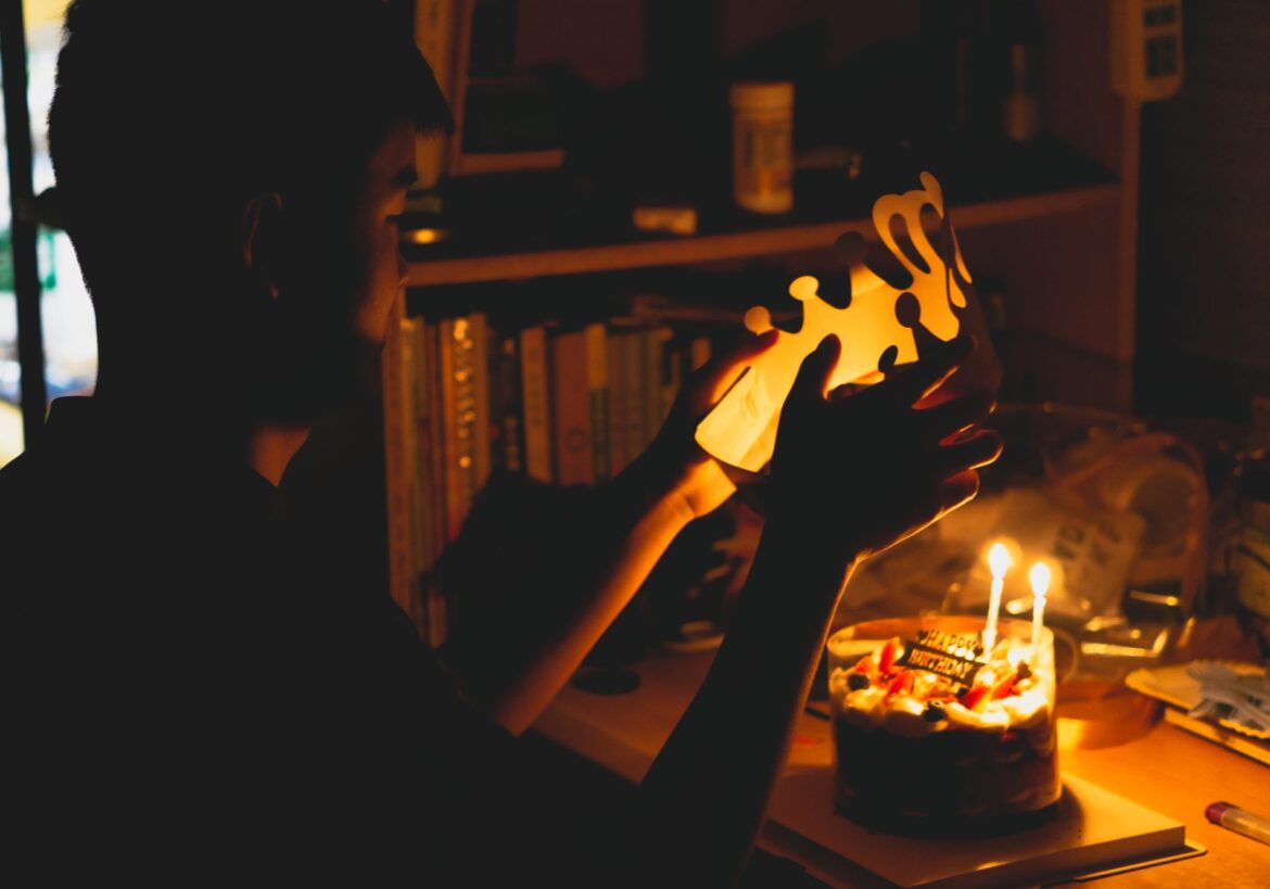 A child prepares to wear a crown beside a lit birthday cake in a warm, cozy indoor setting.