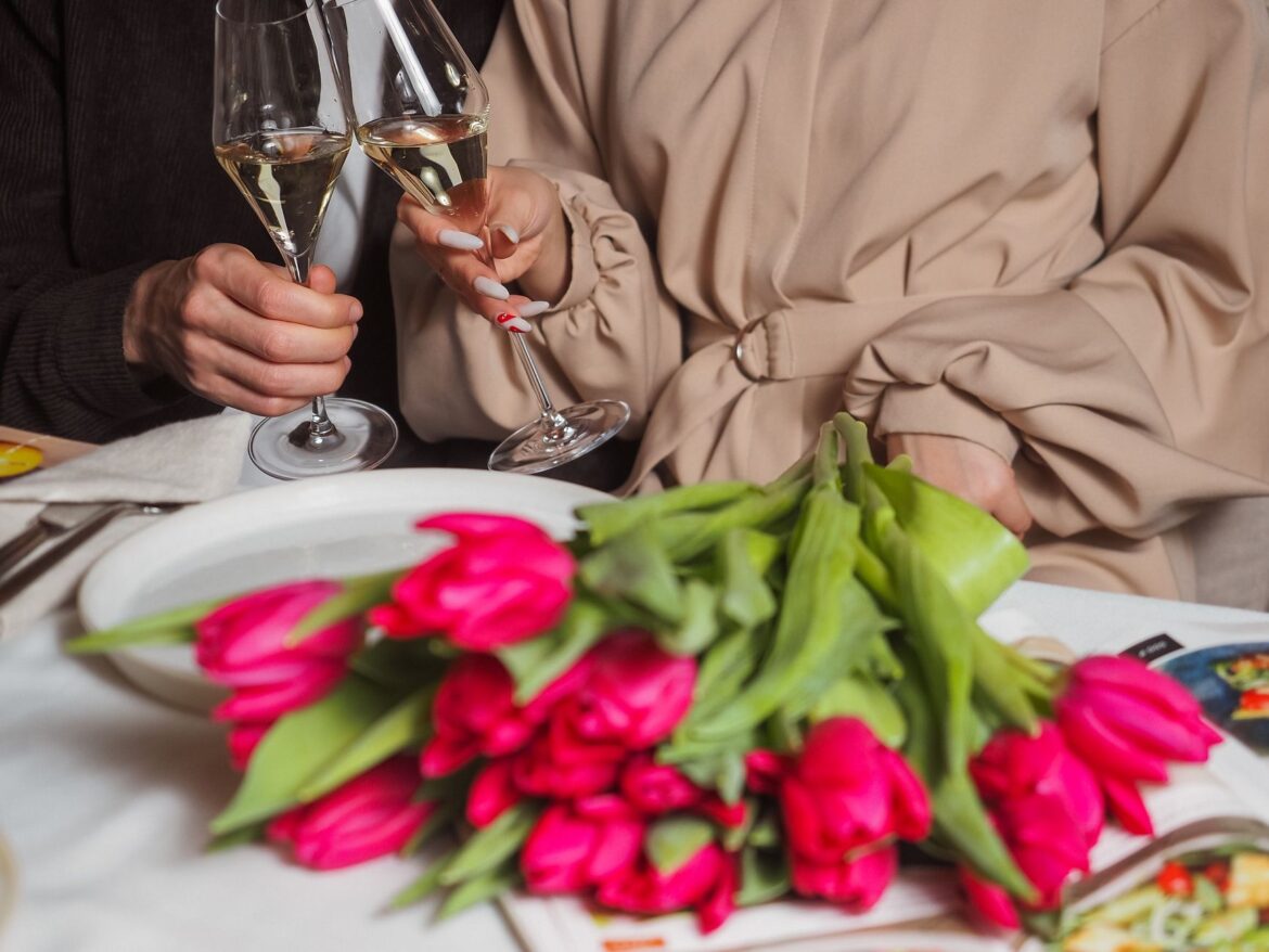 A couple toasting with champagne glasses over a bouquet of pink tulips on a dining table.