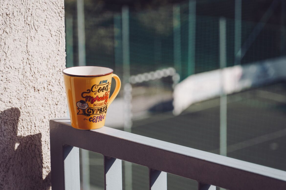 A vibrant yellow coffee mug on a sunlit balcony railing with urban tennis court background.