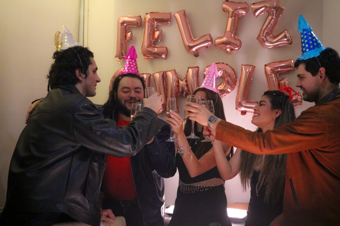Five friends toasting with champagne at a birthday party, wearing colorful hats.