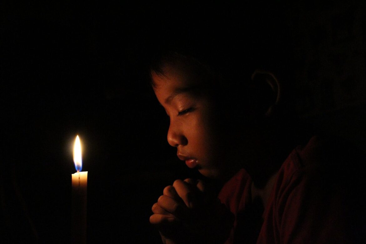 A child prays by candlelight in a dark room, creating a serene, reflective atmosphere.