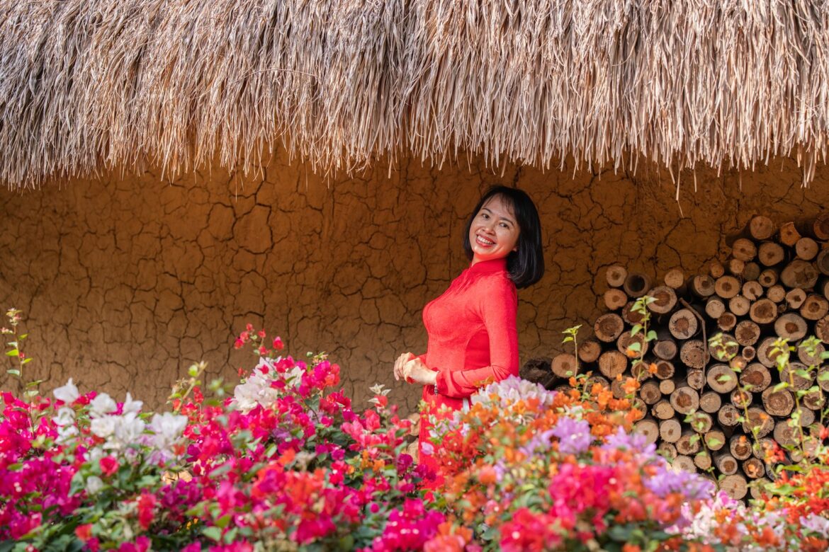 A woman in a vibrant red dress stands smiling among colorful flowers against a rustic backdrop.