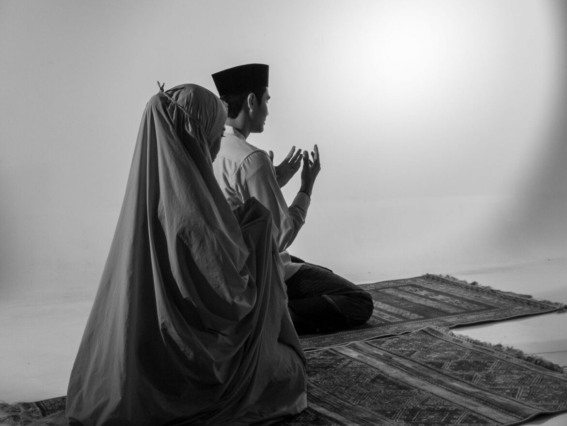 A Muslim couple praying on prayer rugs, captured in a black and white studio setting.