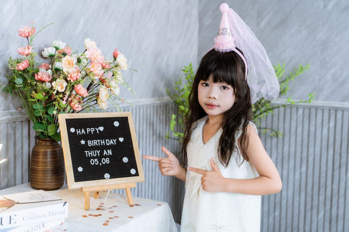 A young girl wearing a birthday hat poses next to a birthday board, celebrating indoors.