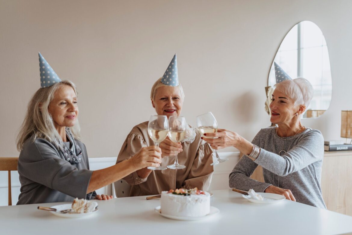 Three senior caucasian women celebrate a birthday with champagne toast and cake indoors.
