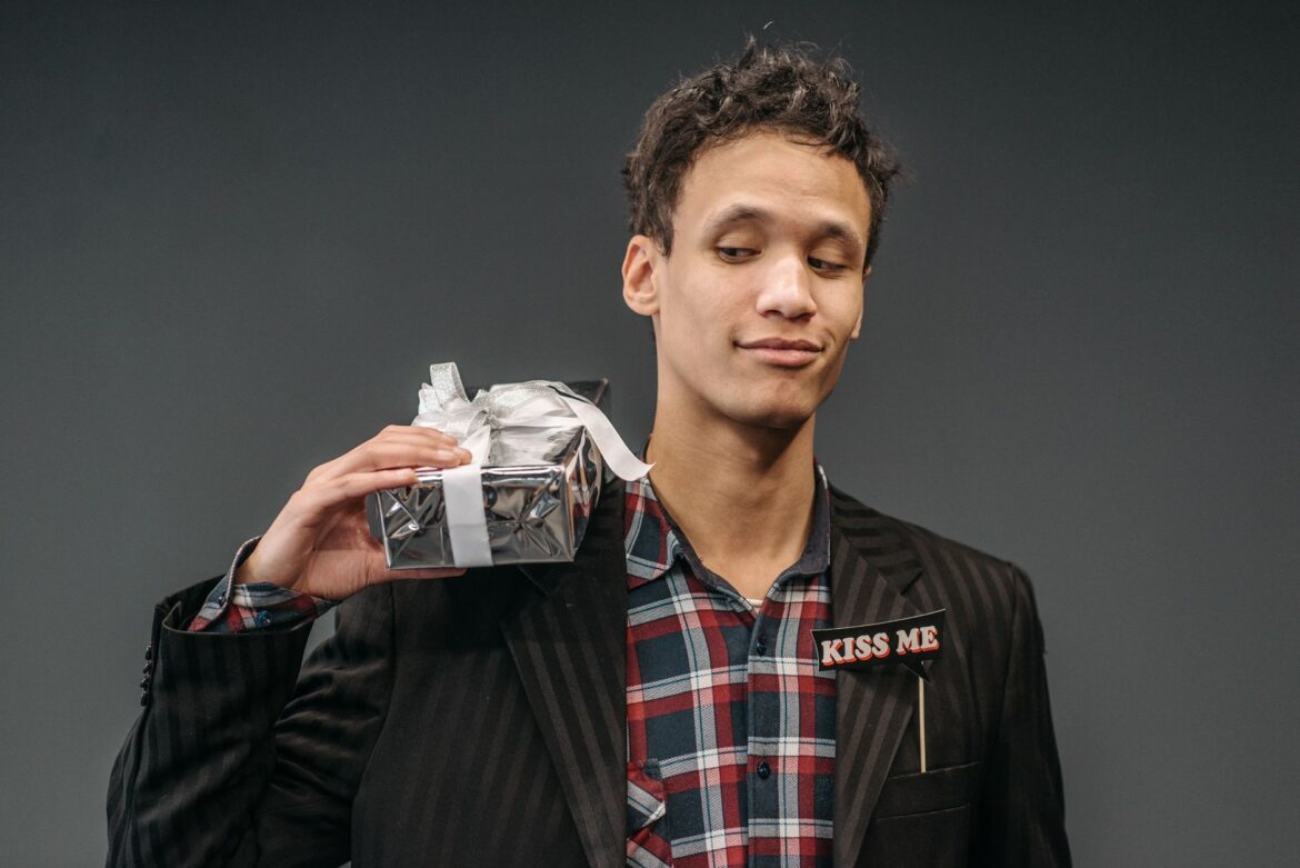 Young man in a plaid shirt and striped suit holds a gift, showcasing a playful expression in a studio setting.