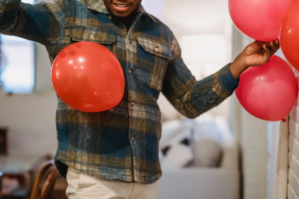 Crop unrecognizable African American male in warm checkered shirt decorating living room with red and pink balloons while preparing for Saint Valentines Day celebration