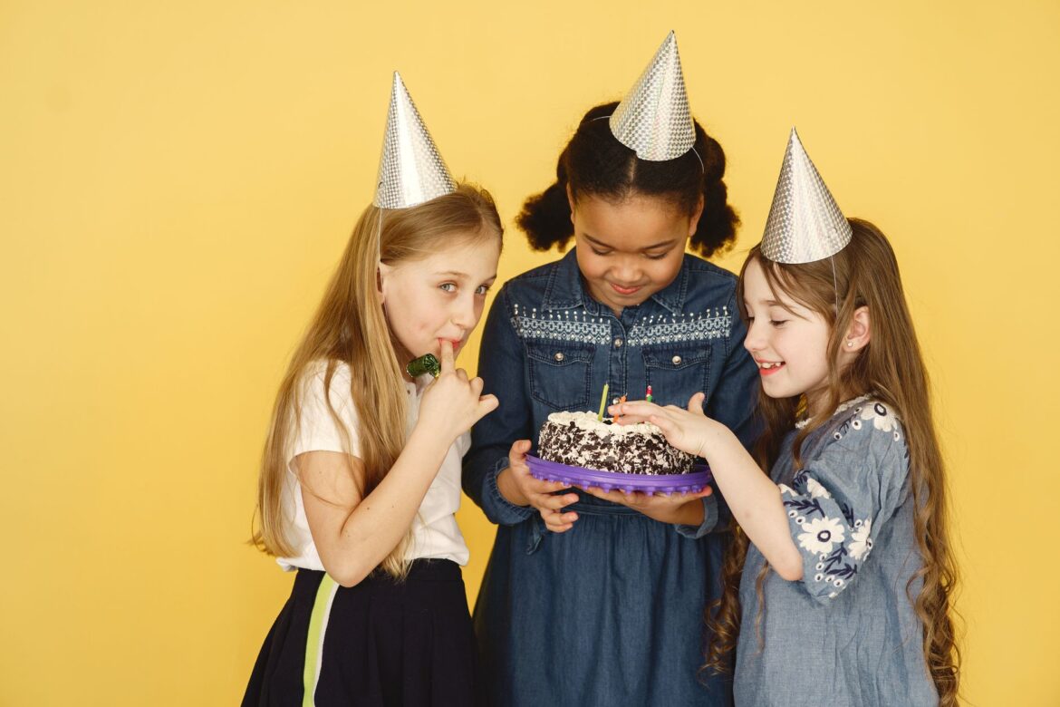 Three children enjoying a birthday party with cake and party hats on a yellow background.
