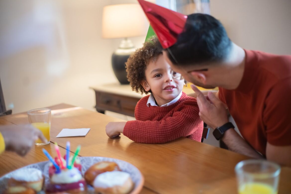 A joyful indoor birthday party scene with a father and son wearing festive hats and enjoying desserts.