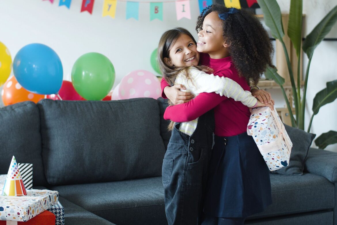 Two girls hugging at a lively birthday party with balloons and gifts indoors.