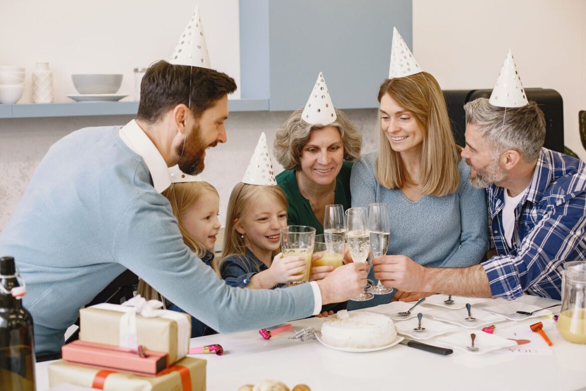 A joyful family toasts at a celebration wearing party hats with a cake and gifts on the table.