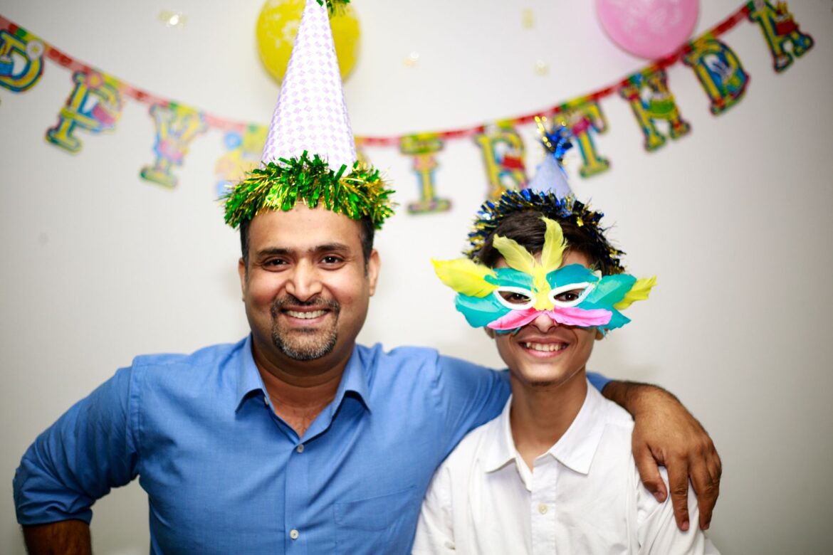 Smiling father and son celebrating a birthday with party hats and decorations indoors.