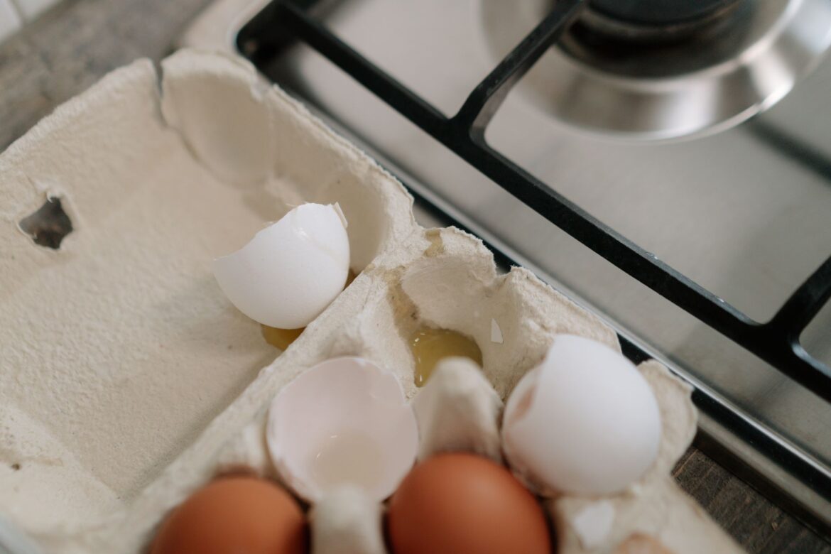 Egg carton with brown and white eggs and broken shells on kitchen stovetop.