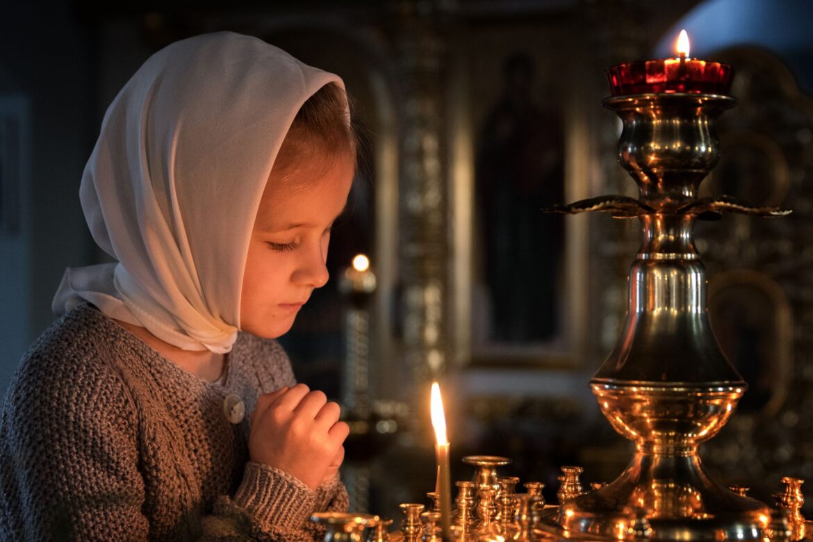 A young girl in a headscarf praying with candles in a church setting.