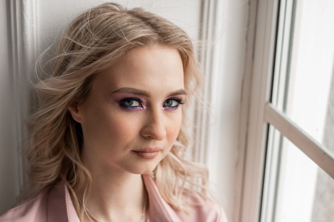 Close-up portrait of a blonde woman by a window with soft natural lighting.