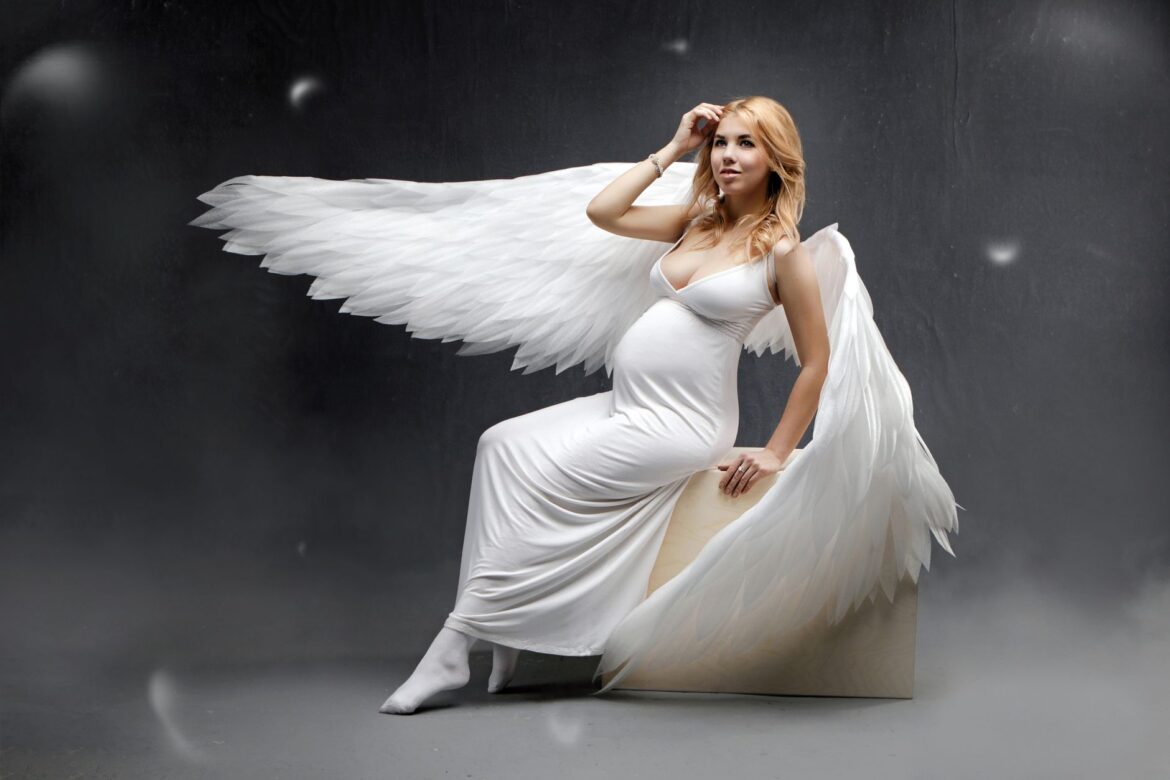 Pregnant woman in an angel costume with wings, seated on a cube in a studio.
