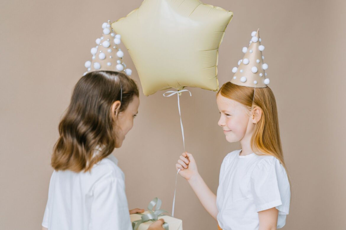 Two girls at a birthday party wearing hats, exchanging a gift and balloon.
