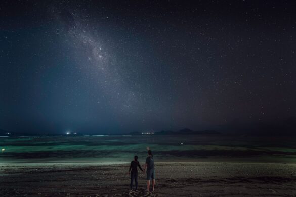 Romantic couple holding hands while observing stars on a beach at night, showcasing love and serenity.
