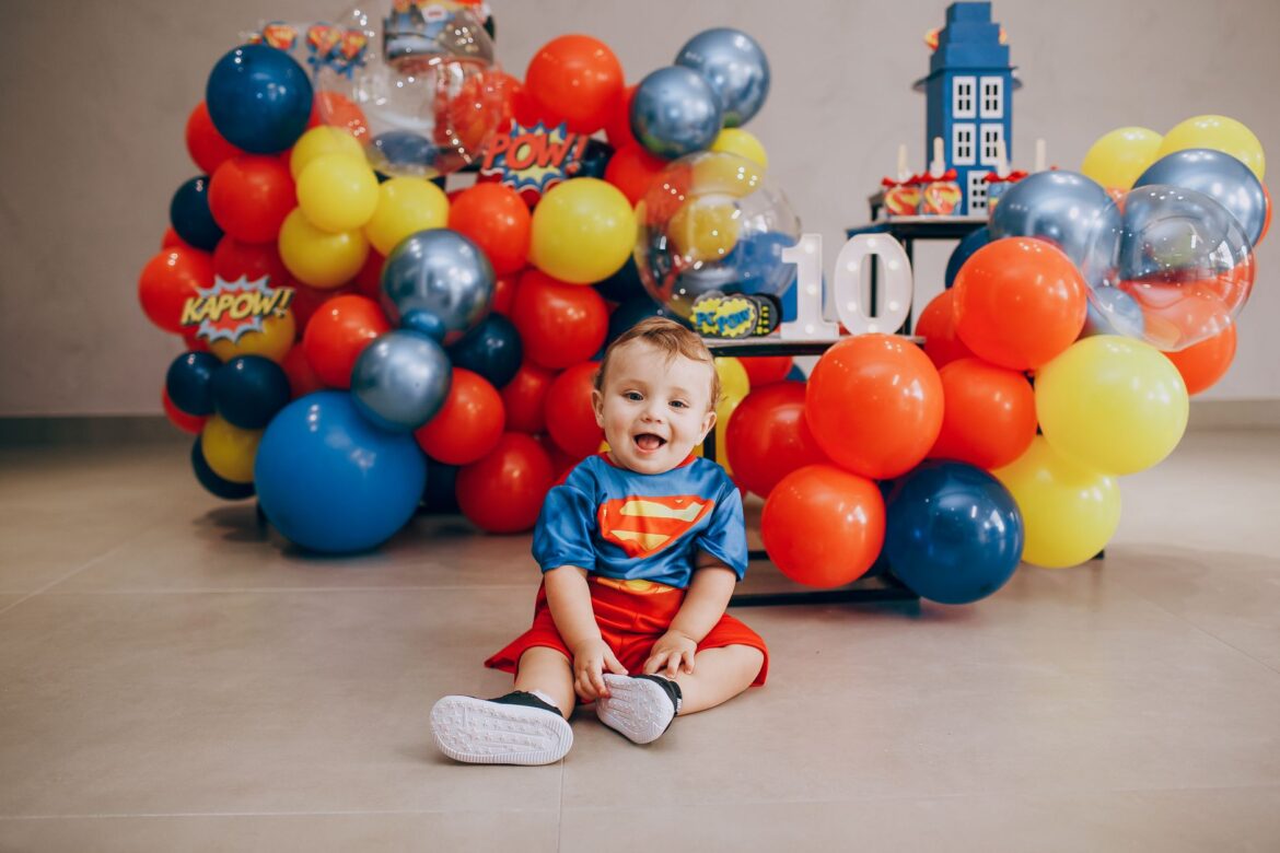 Cute toddler in Superman costume amidst colorful balloons celebrating birthday indoors.