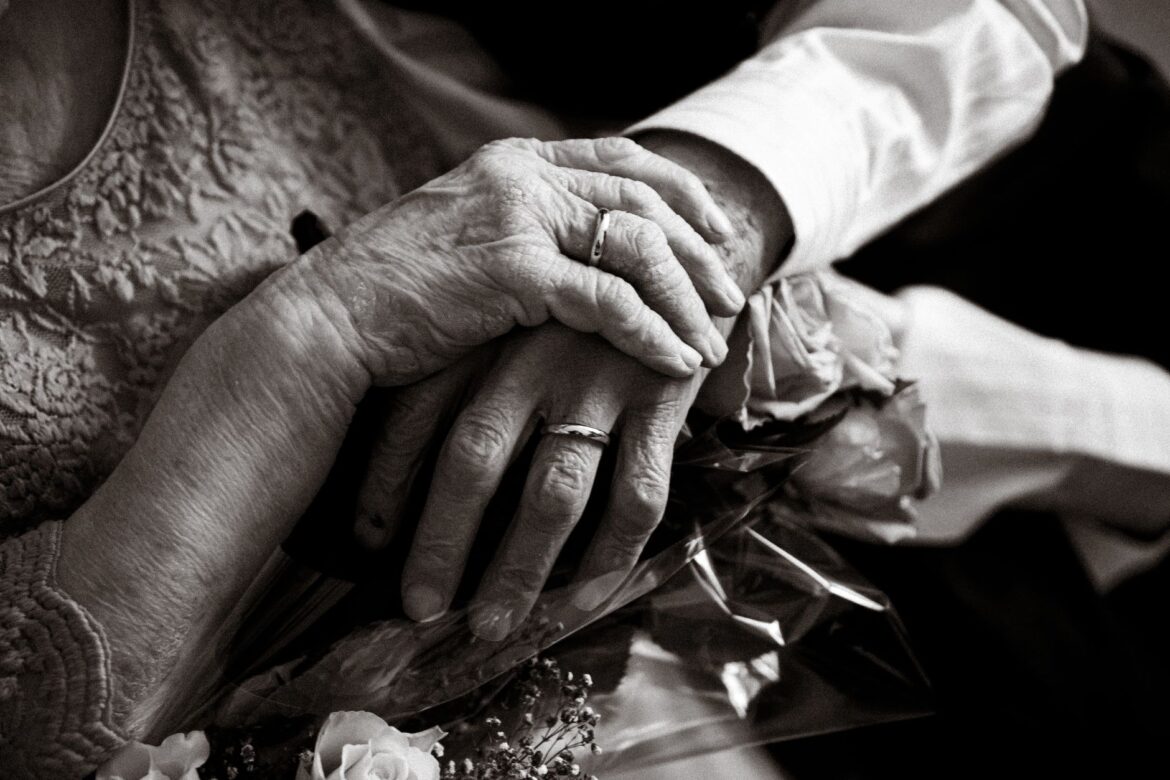 Black and white photo of an elderly couple holding hands with wedding rings, symbolizing love and companionship.