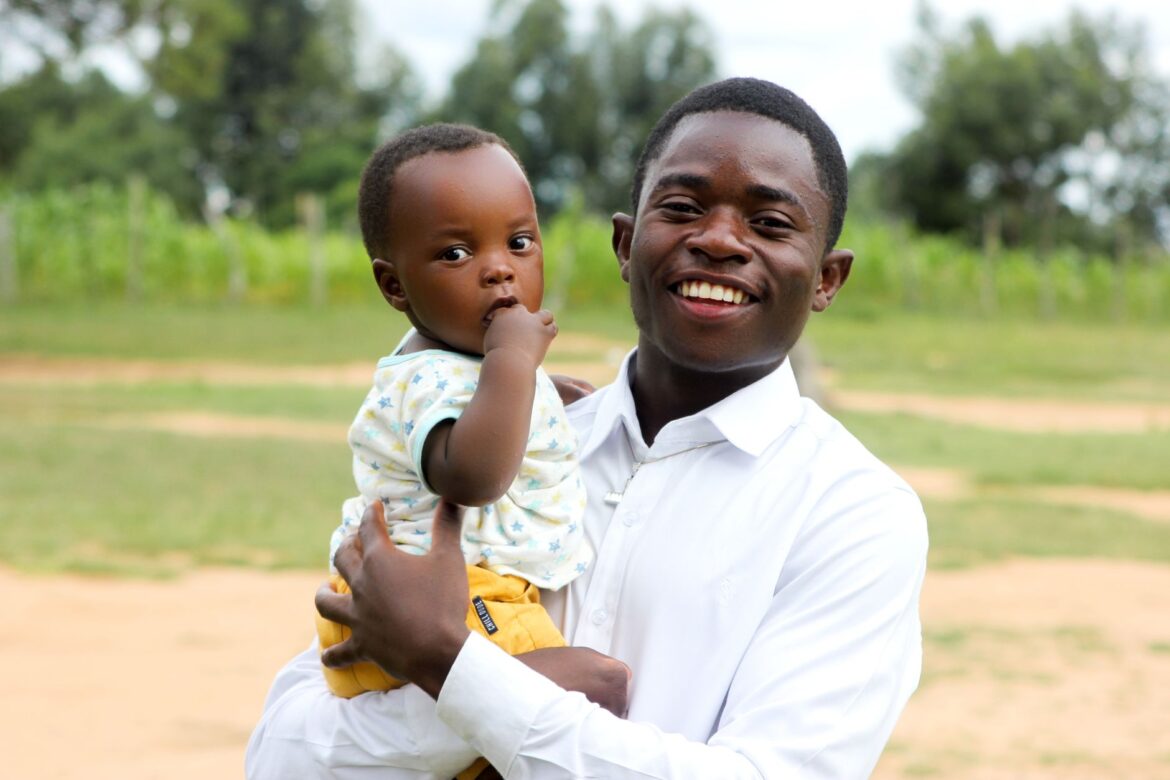 A joyful young man holds his baby outdoors in a park, both smiling at the camera.