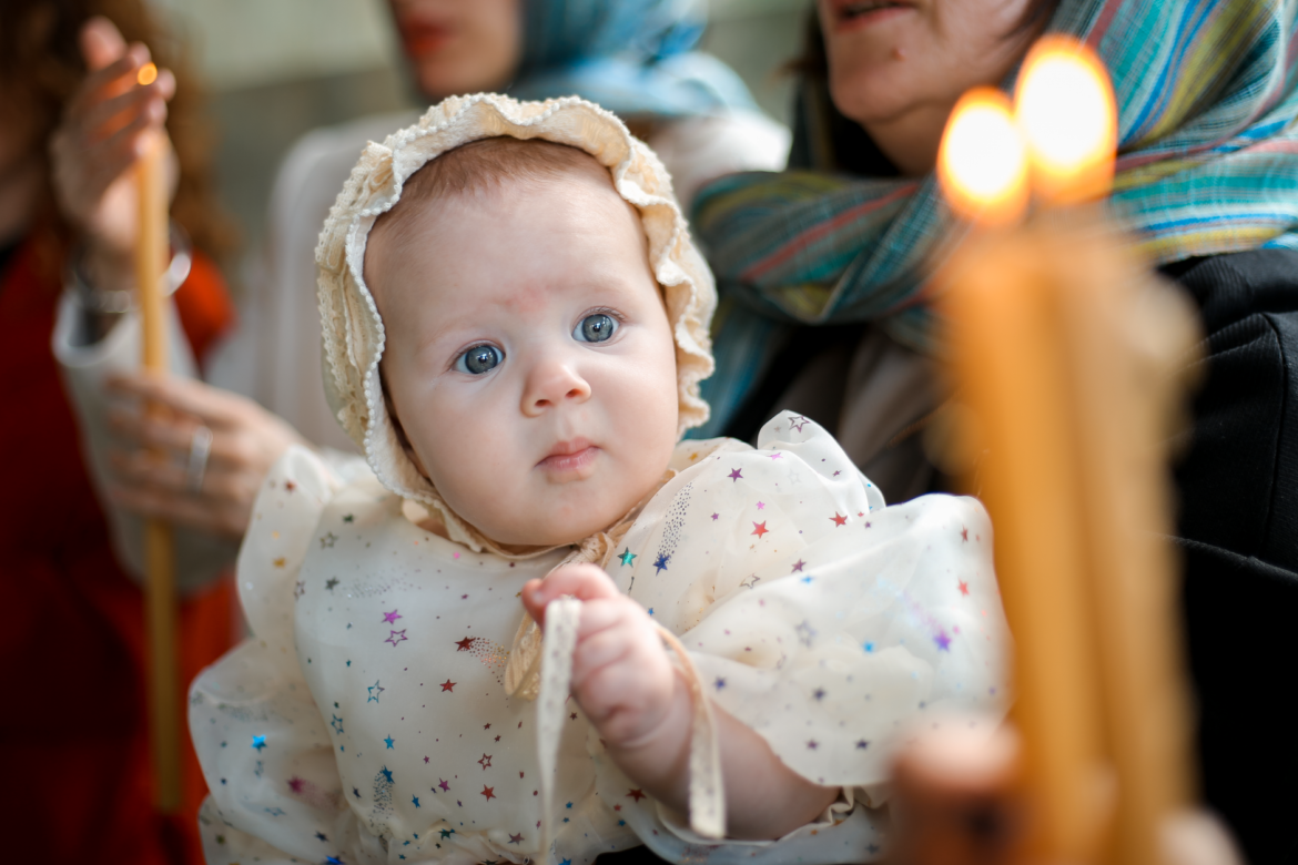 A baby in a bonnet during a baptism ceremony, surrounded by candles and family.