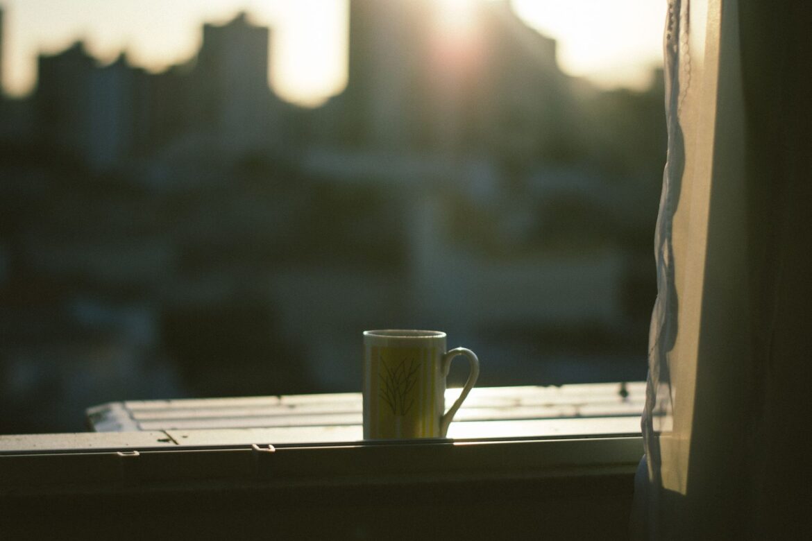 A mug sits on a windowsill with the city skyline and sunrise in the background.
