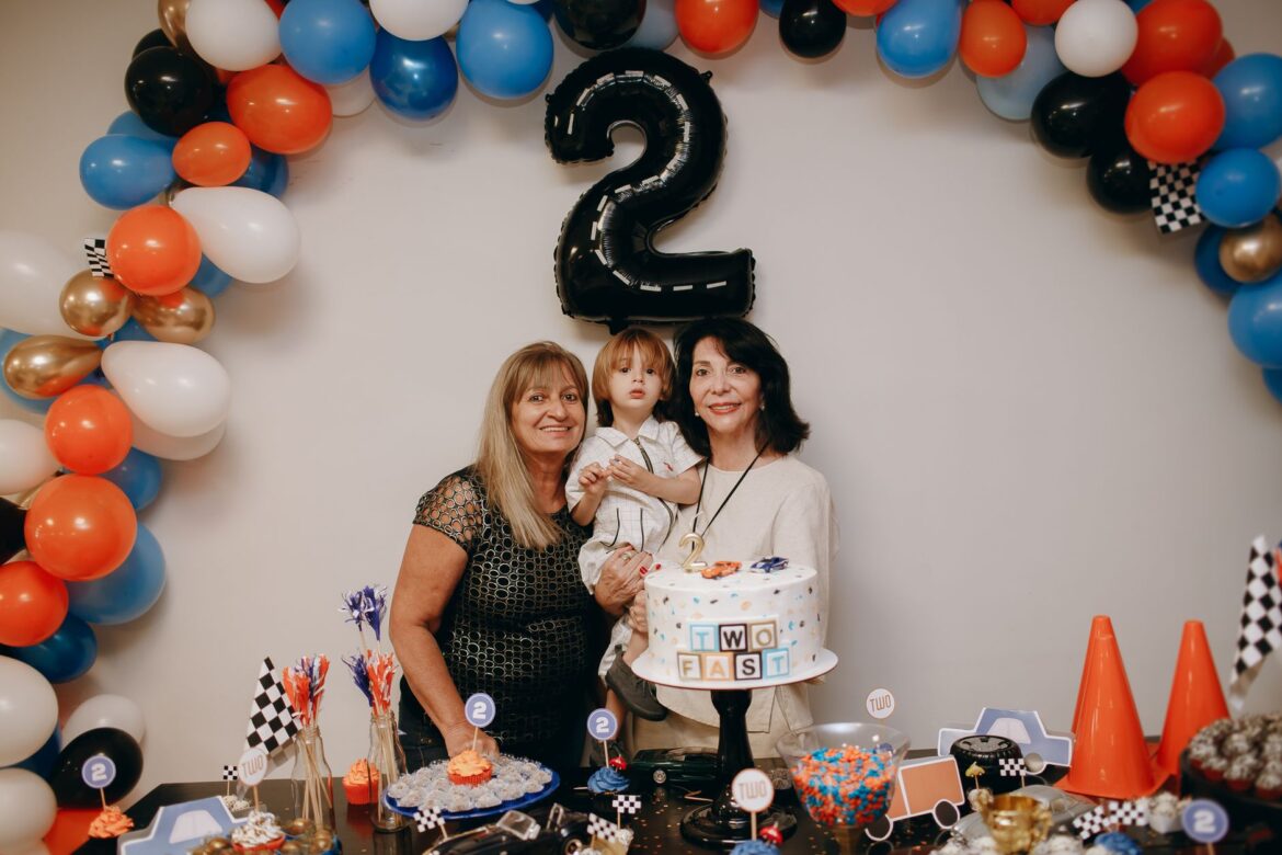 A family celebrates a child's second birthday with balloons and a themed cake.