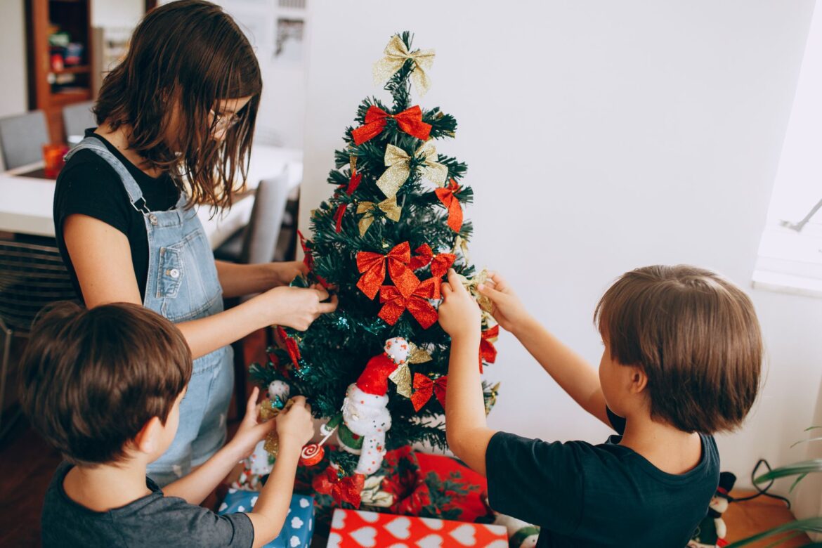 Three kids decorating a Christmas tree with bows and ornaments indoors, spreading holiday cheer.