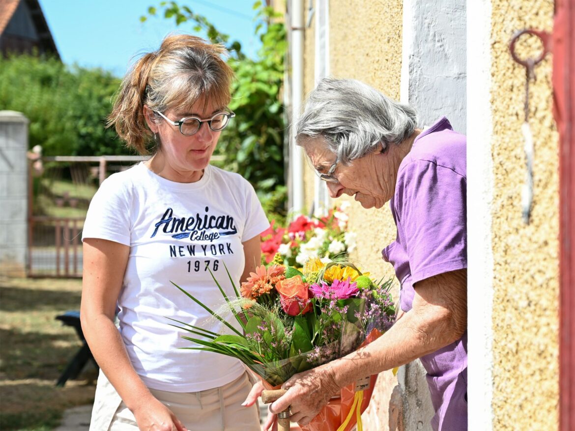 A woman gifts a vibrant bouquet to an elder outside a French house.