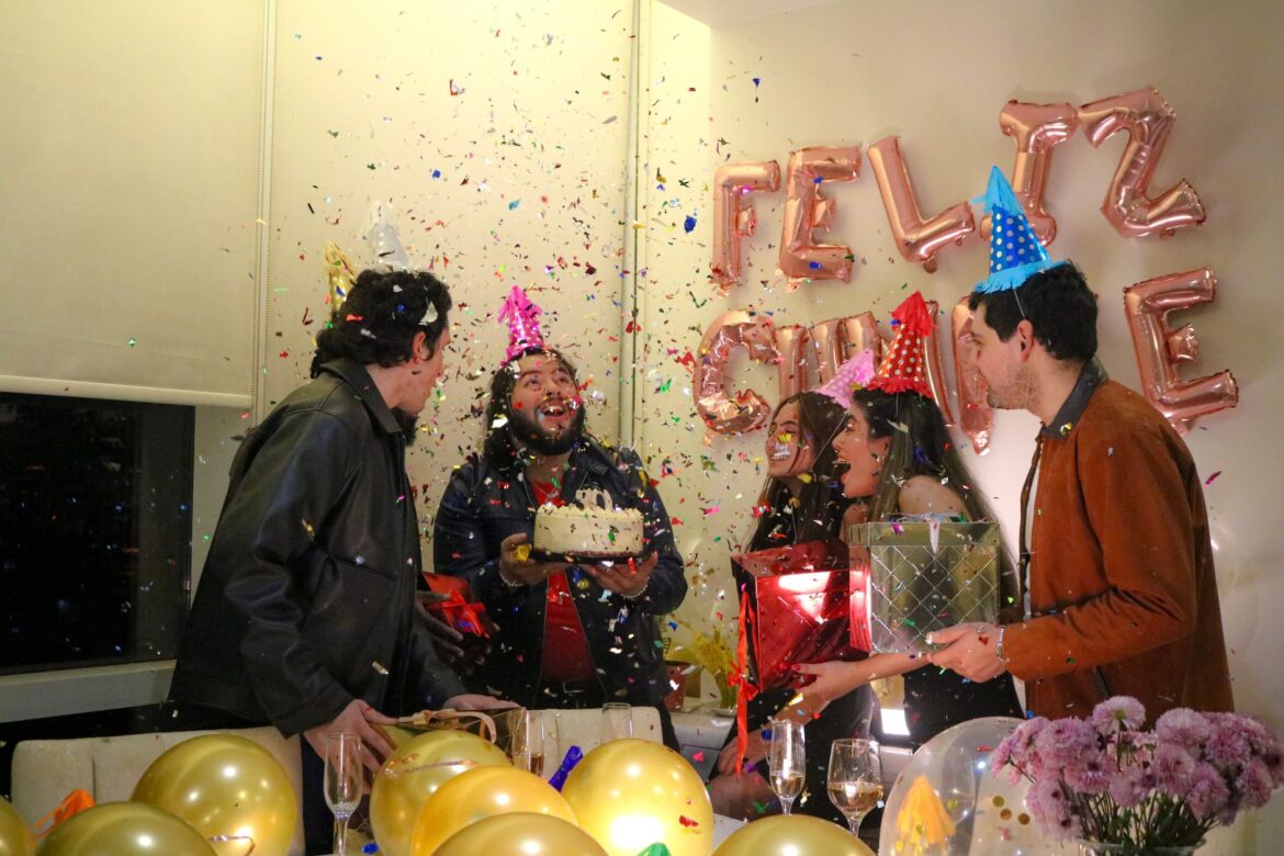 Group celebrating a birthday with cake, balloons, and confetti indoors.
