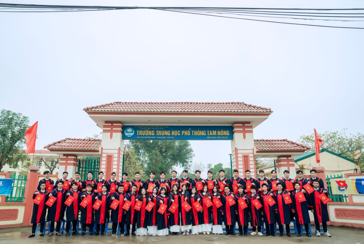 Large group of students in graduation robes posing in front of Tam Nong High School gate.