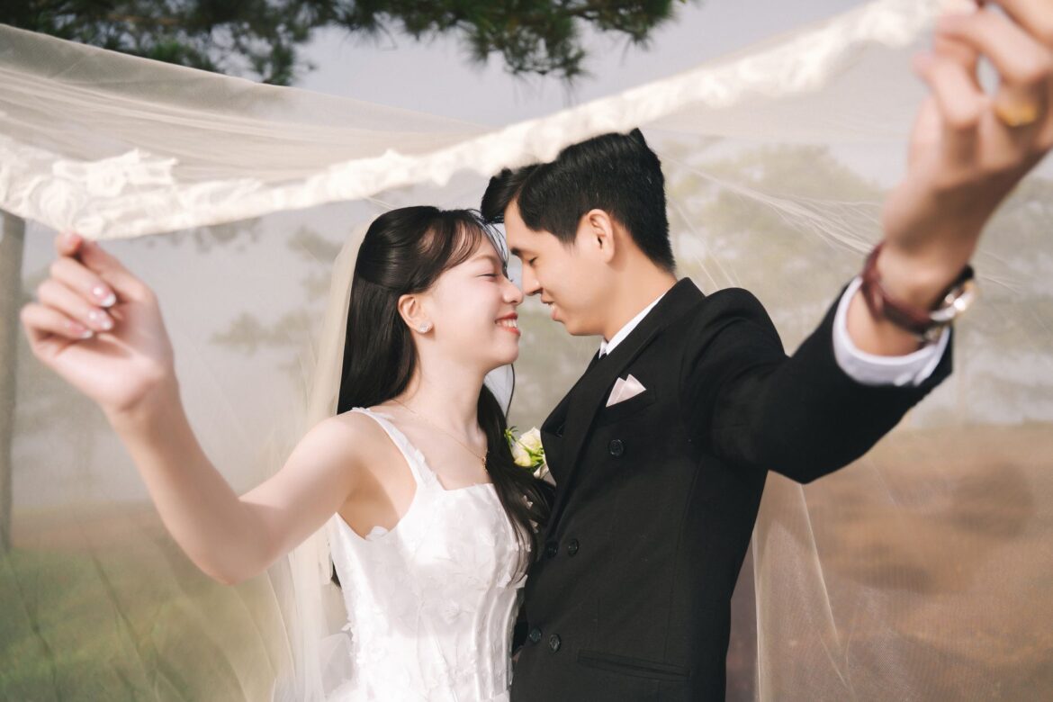 A joyful bride and groom embrace under a veil in an outdoor wedding scene.