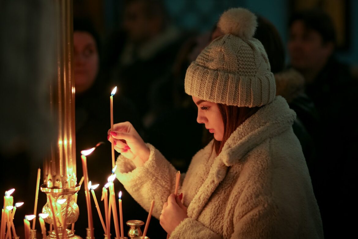 A woman in winter attire lights a candle during a religious ceremony indoors.