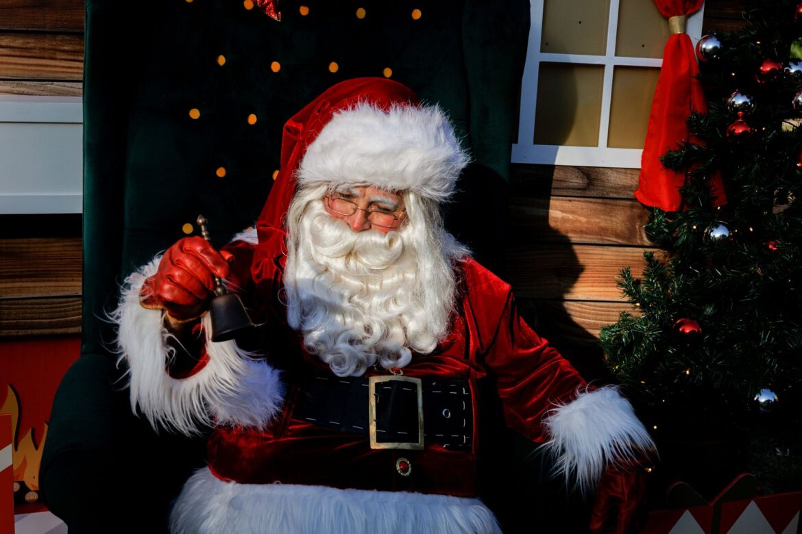 Festive Santa Claus ringing a bell next to a decorated Christmas tree indoors.