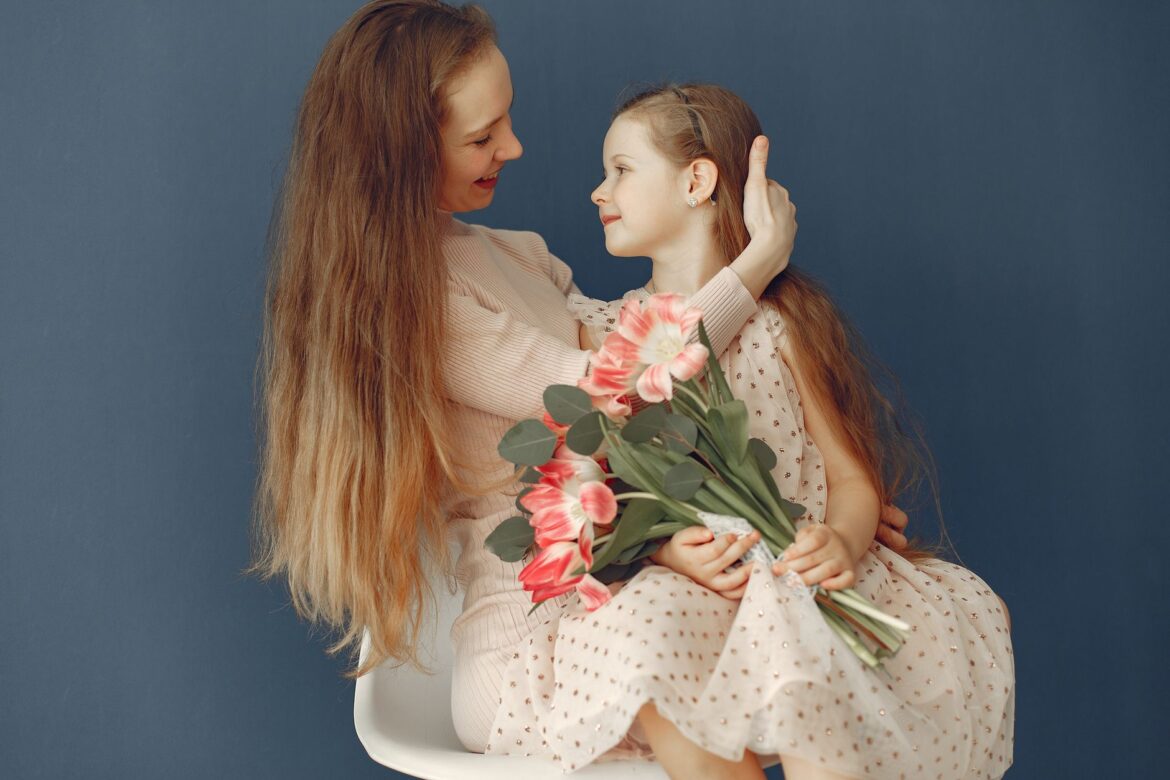 Side view of red haired lady in dress sitting on chair and cuddling cute girl with flowers while mom and daughter looking at each other