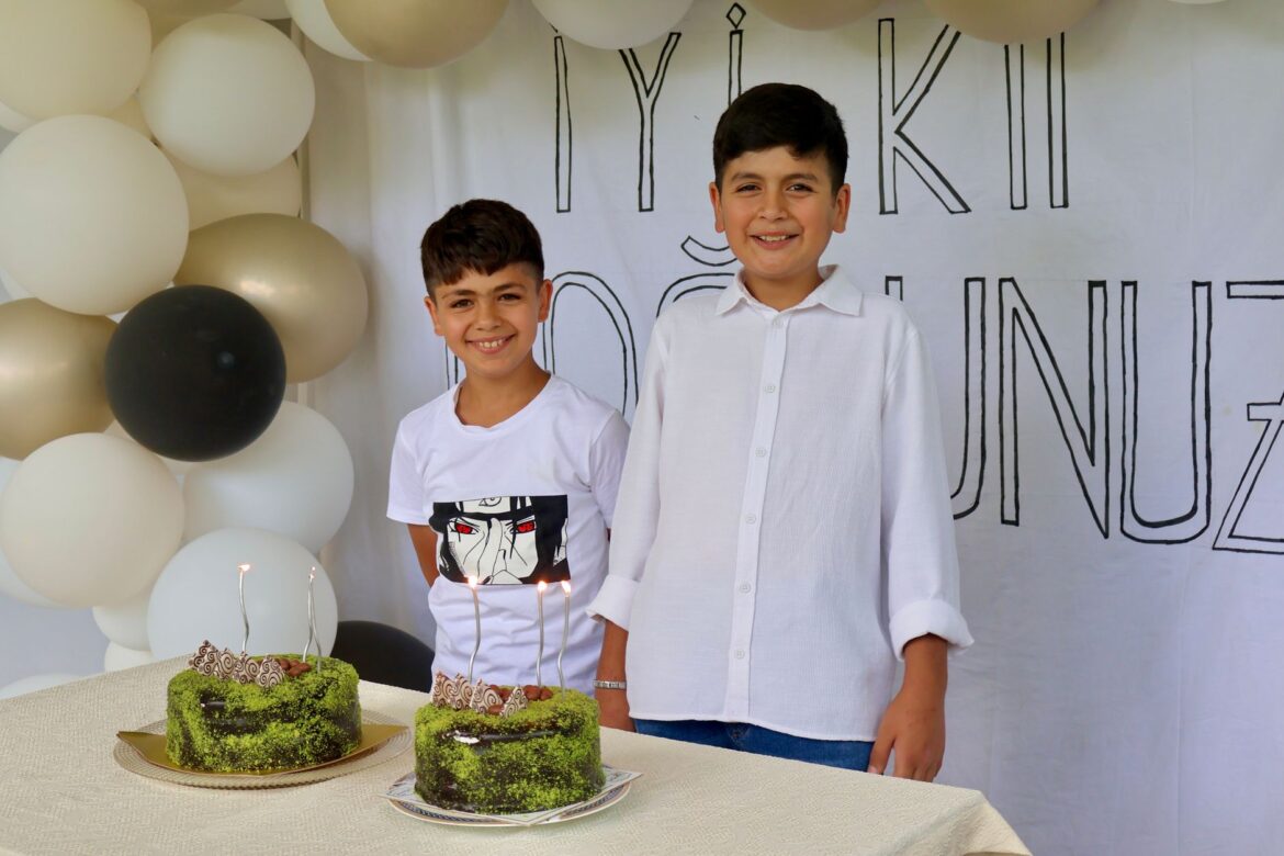 Two boys smiling at a birthday party with cakes and decorative balloons.