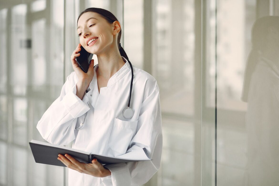 Smiling ethnic female doctor in medical uniform with stethoscope and notebook talking on smartphone while standing in glass hospital corridor