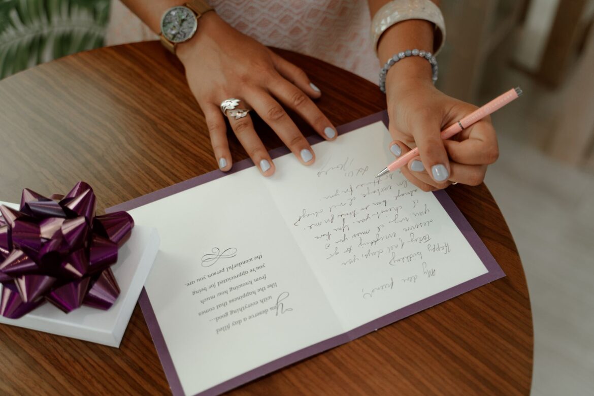 A woman writes a message inside a greeting card on a wooden table with a gift beside.