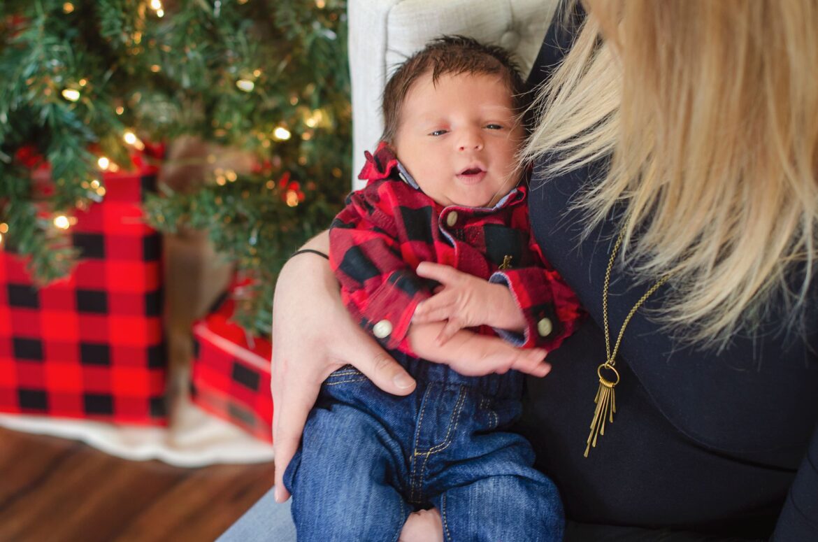 A mother cradles her newborn in a cozy Christmas setting with a decorated tree and gifts.