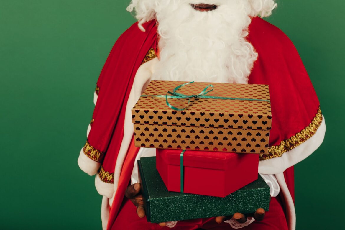 Santa Claus holding stacked gift boxes against a vibrant green background, ready for Christmas celebrations.