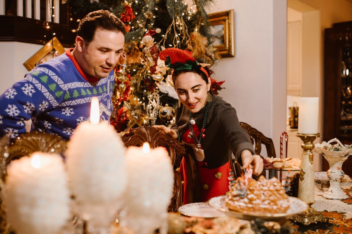 Happy adult multiethnic couple serving table while standing near decorated fir tree during Christmas celebration at home