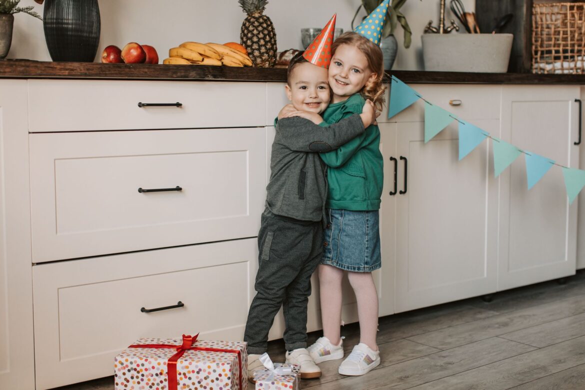 Two children wearing party hats hugging at a birthday celebration in a kitchen setting.