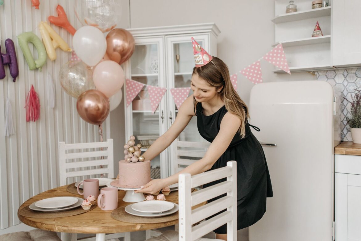 A woman arranges a birthday cake on a table with balloons and decorations in a festive indoor setting.