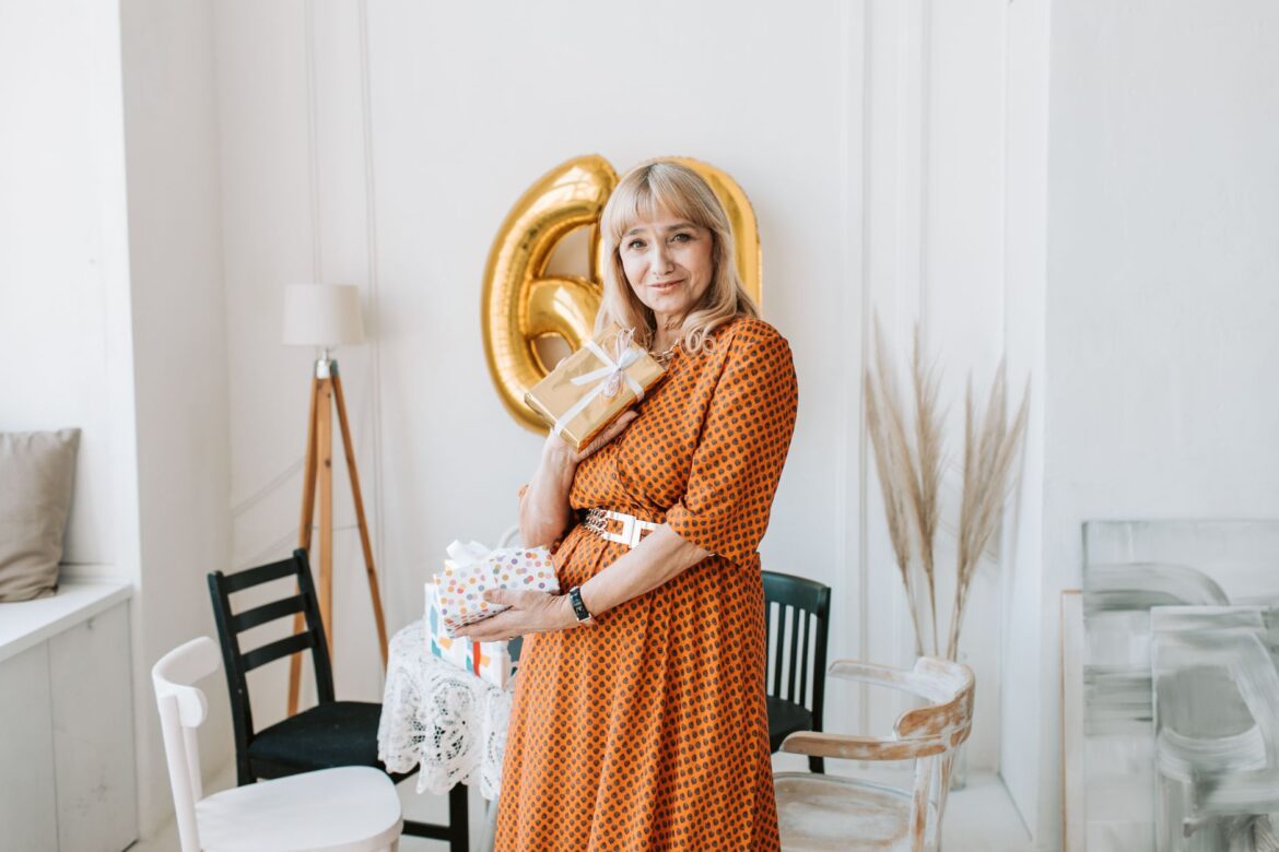 Elderly woman in orange dress holding gifts and smiling, celebrating birthday indoors.