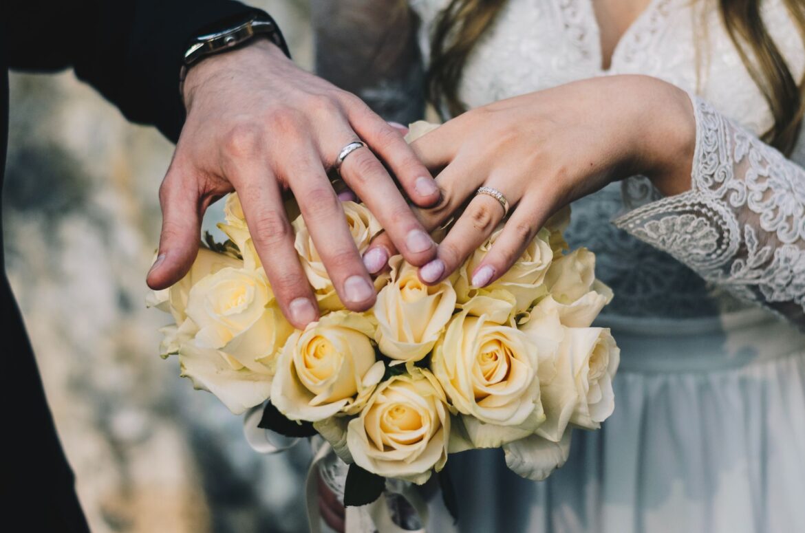 Close-up of a couple's hands displaying wedding rings above a bouquet of yellow roses.