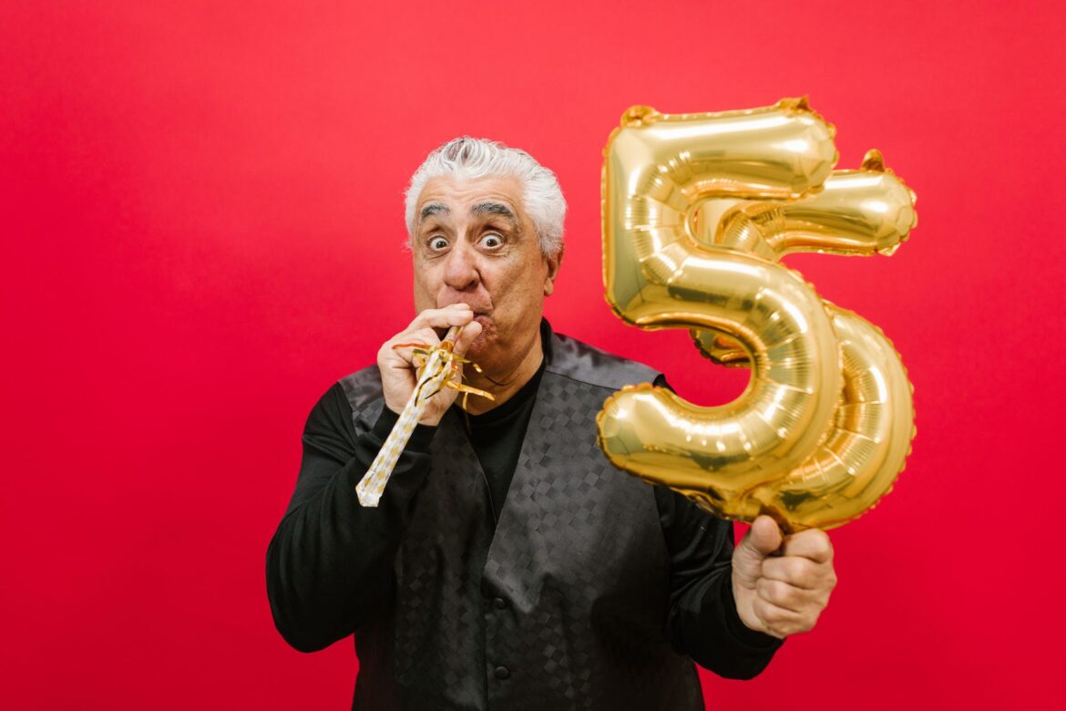 Senior man with a party horn and gold balloon, celebrating in a studio with a vibrant red background.