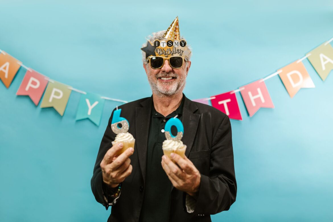 Happy senior man celebrating 60th birthday holding cupcakes with candles.