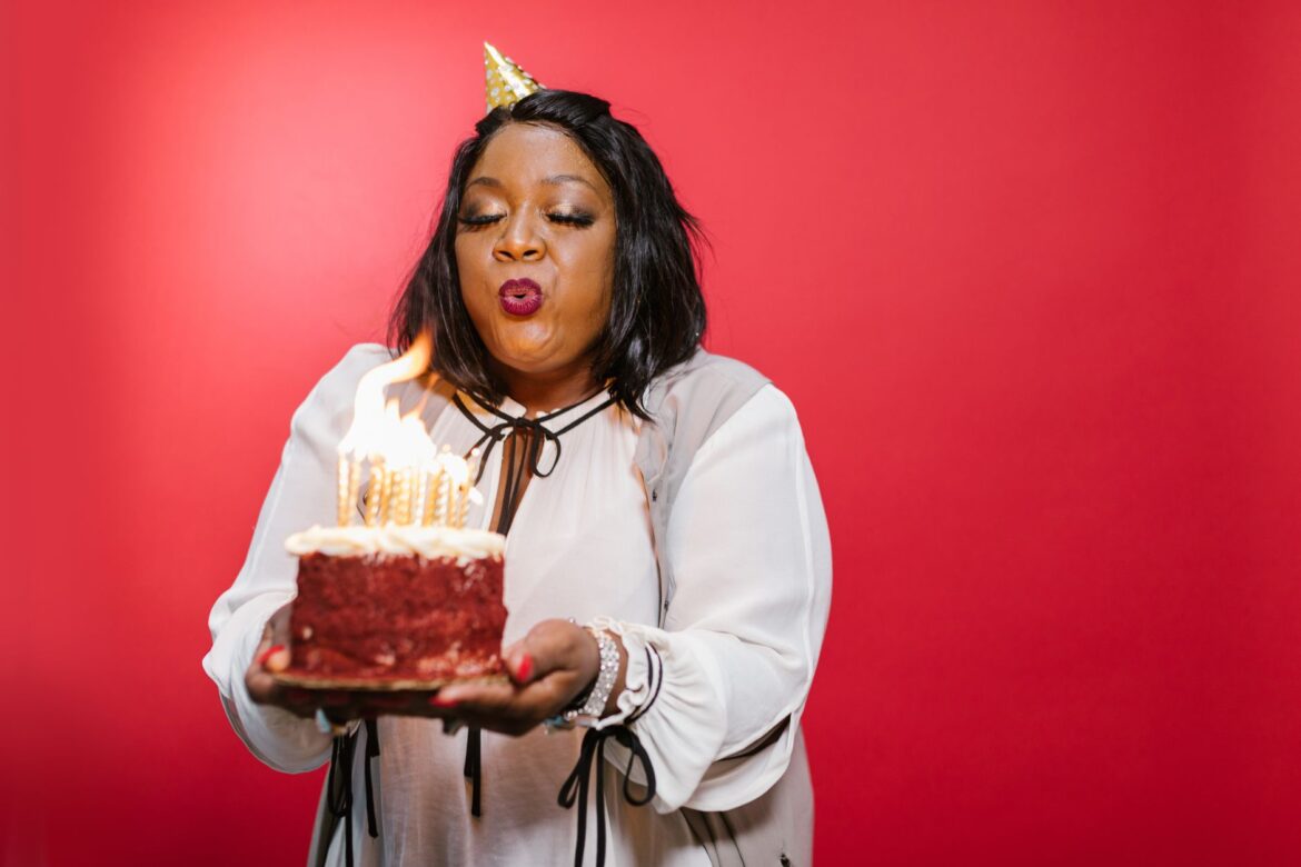 A black adult woman joyfully blowing out birthday candles on a red velvet cake against a red backdrop.